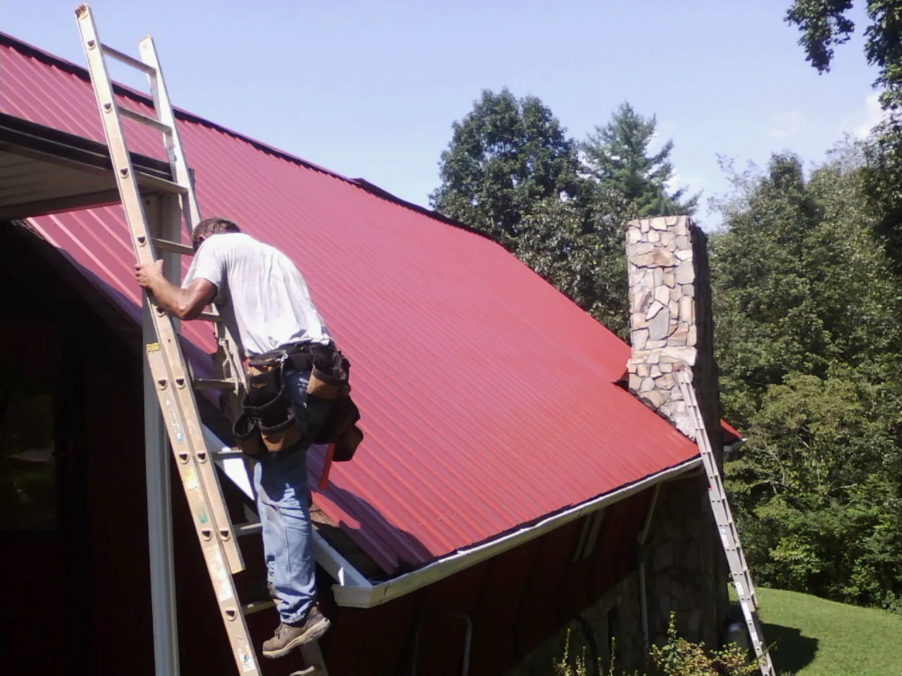 Roofer on a ladder beside a red metal roof and stone chimney — Soffit & Fascia Repair by Roofing Brothers in Calhoun Falls